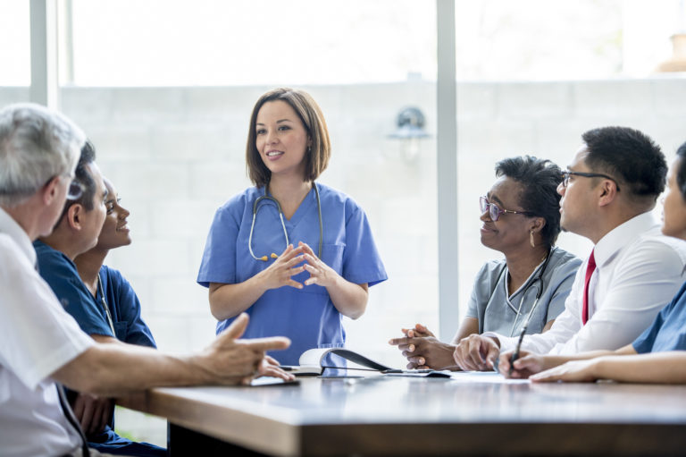 A multi-ethnic group of medical staff are indoors in a hospital. They are wearing medical clothing. A Caucasian female doctor is giving a presentation to the others.